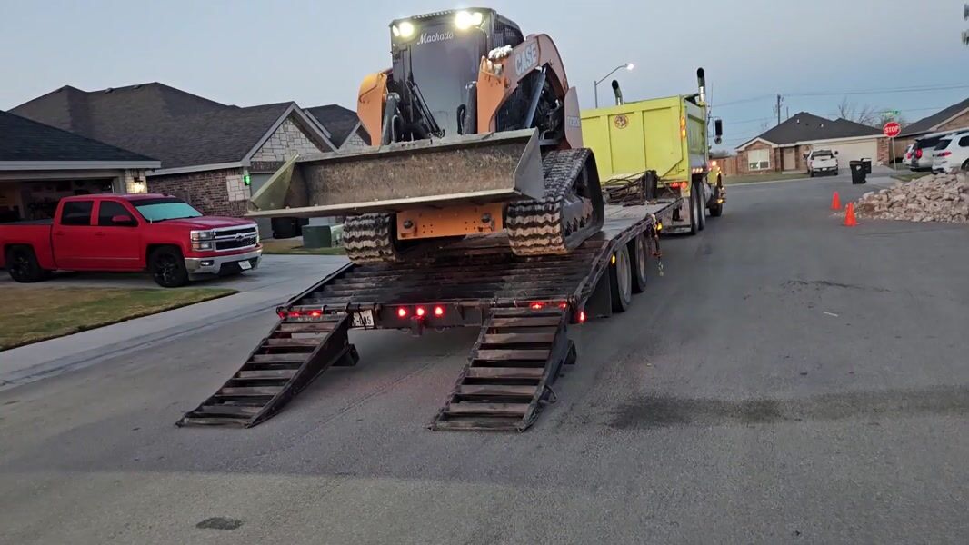Skid steer being unloaded from a trailer on a residential street in Odessa, Texas, with a cleanup pile at the curb.